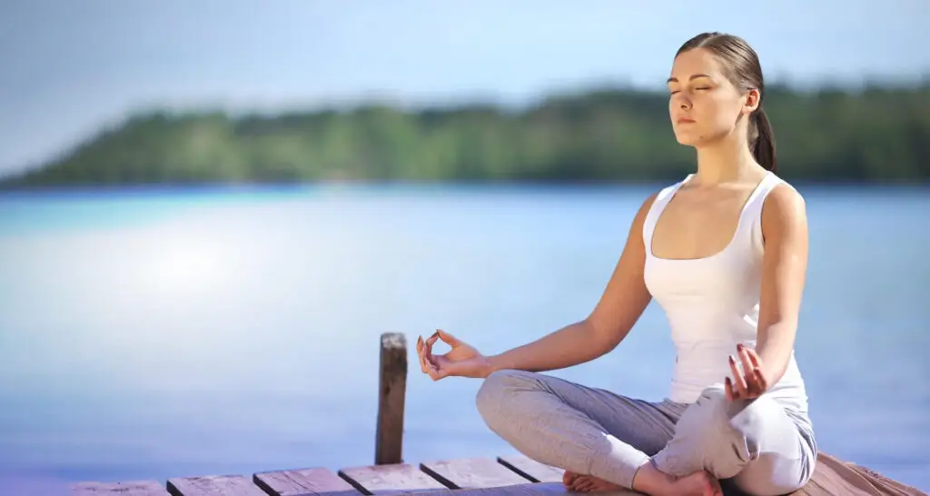 Woman meditating by a tranquil lake, promoting mindfulness and stress reduction techniques for improved mental clarity and emotional stability.