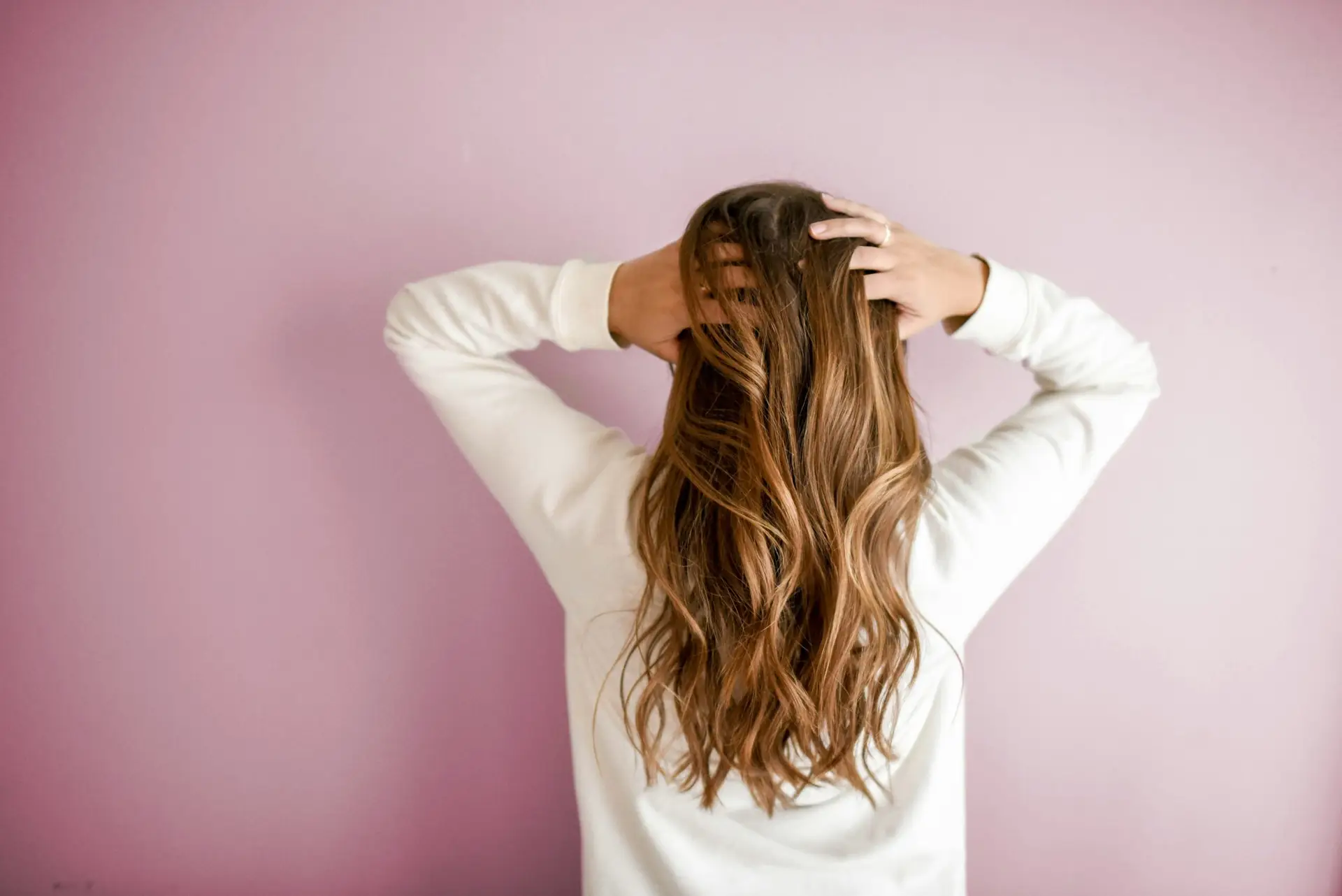 Woman with long, wavy hair touching her scalp against a pink background, representing hair restoration and wellness for men's health products by Prime Vitality Total Wellness and Medical Spa.