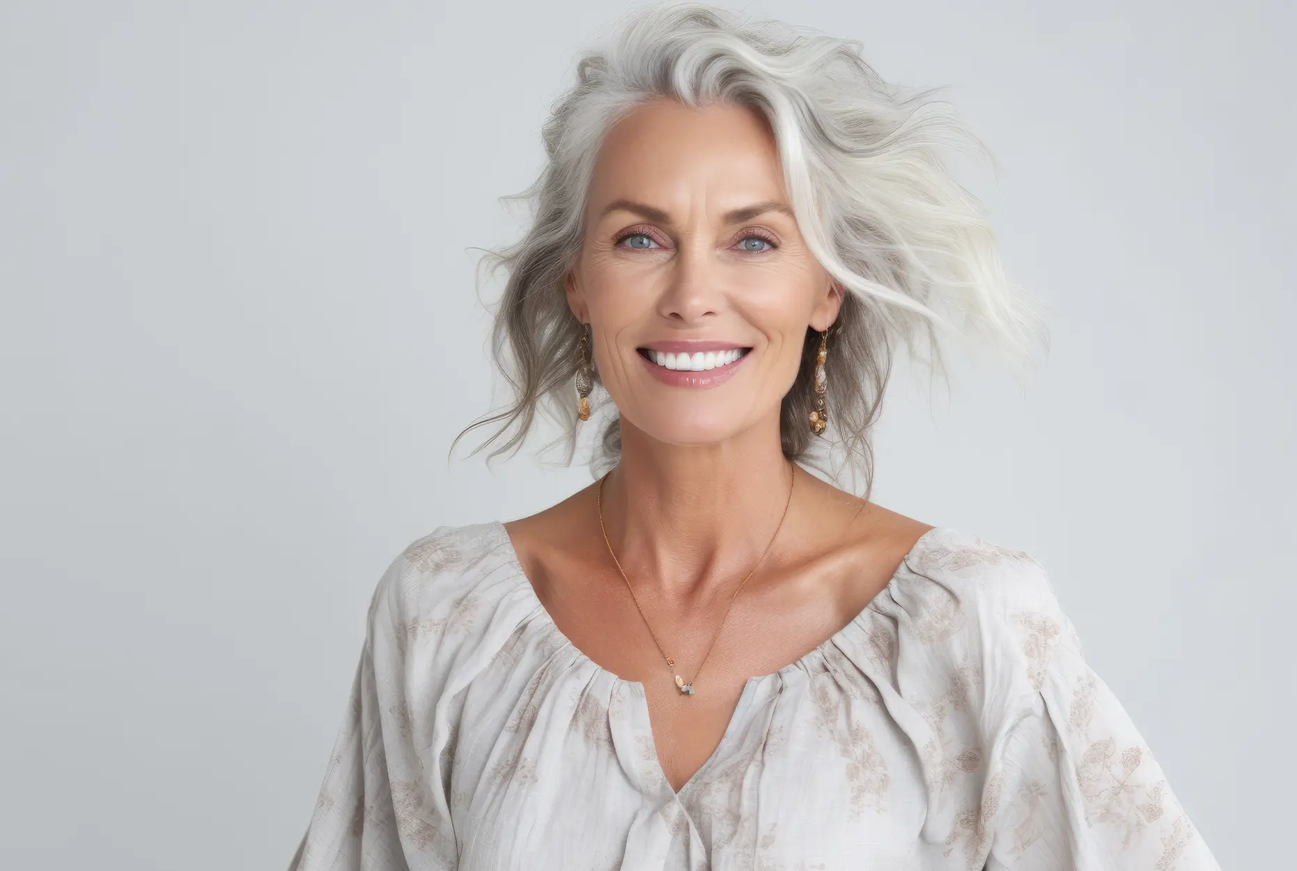Smiling mature woman with silver hair and natural makeup, wearing a light blouse and gold jewelry, representing beauty and vitality in aesthetic treatments.